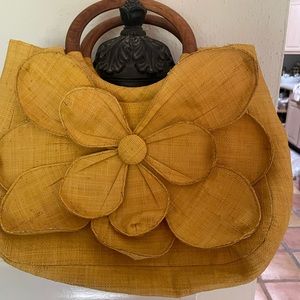 Golden yellow handbag w wooden handle has a flower attached to front of purse.